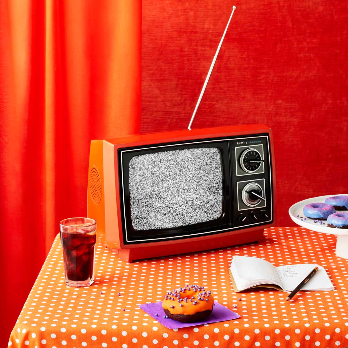 Photograph of a vibrant retro-style kitchen with an Admiral black & white television, donut, notebook, and glass of Coke.